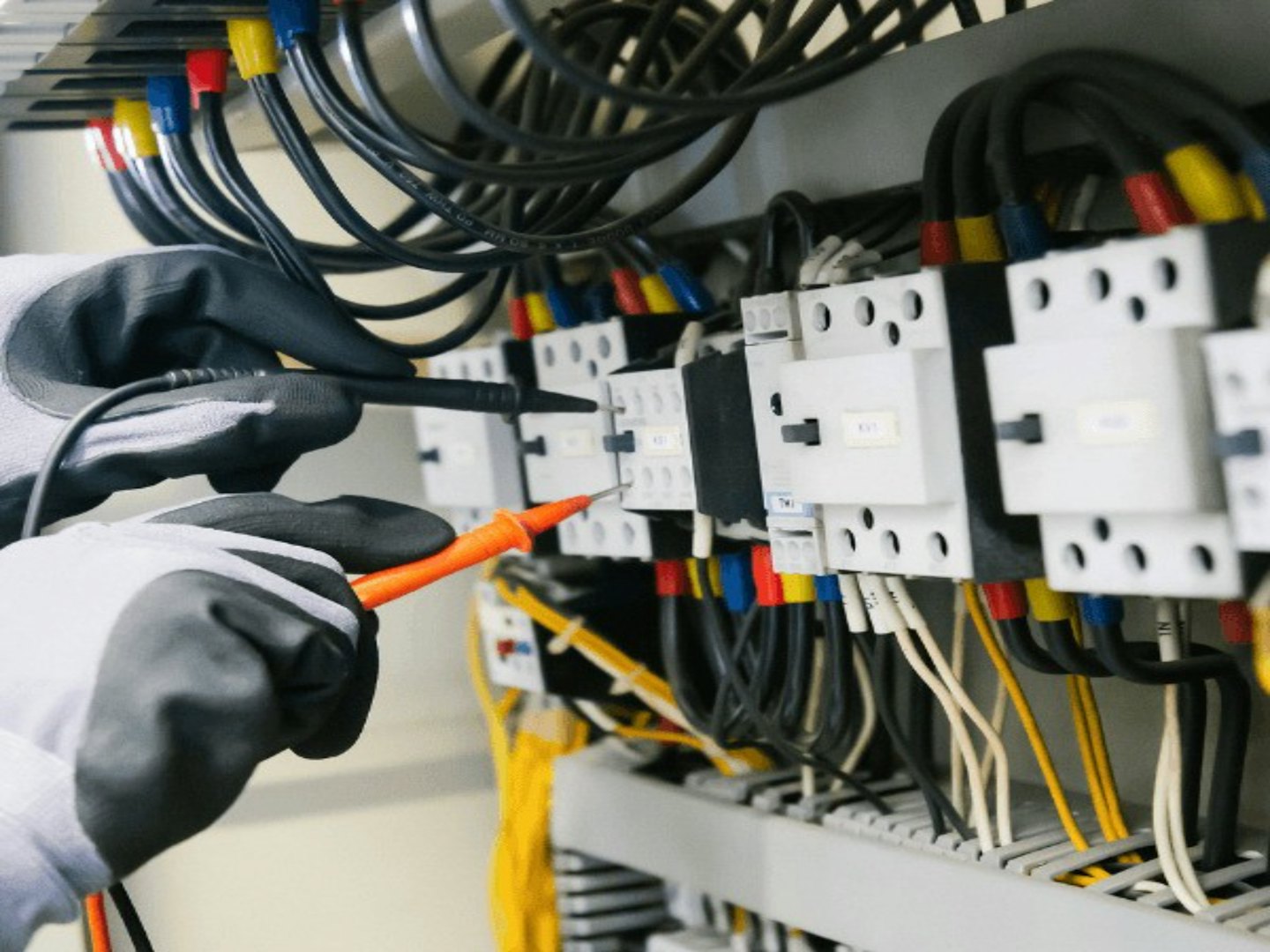 Technician's gloved hands test electrical panel with multimeter.